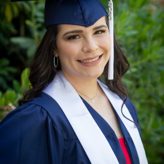 woman with long black hair wearing blue cap and gown and white stole smiling